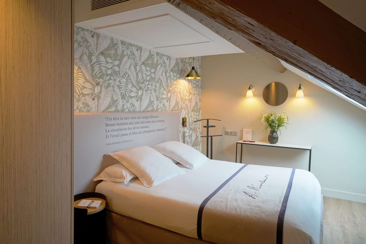 A neatly made bed with a decorative headboard, a bedside table with a vase of flowers, and a wall with a floral wallpaper and a quote.