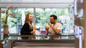 Una pareja sentada en una mesa de vidrio dentro de un edificio moderno con ventanas grandes y vistas al exterior, lleno de vegetación.