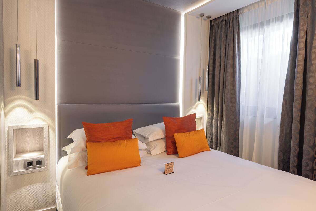 Habitación de hotel con una cama grande, dos almohadas naranjas, una mesita de noche con lámpara y una ventana con cortinas.