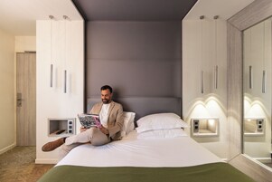 Un uomo seduto su un letto in una camera d'albergo moderna, con armadi a muro e comodini.