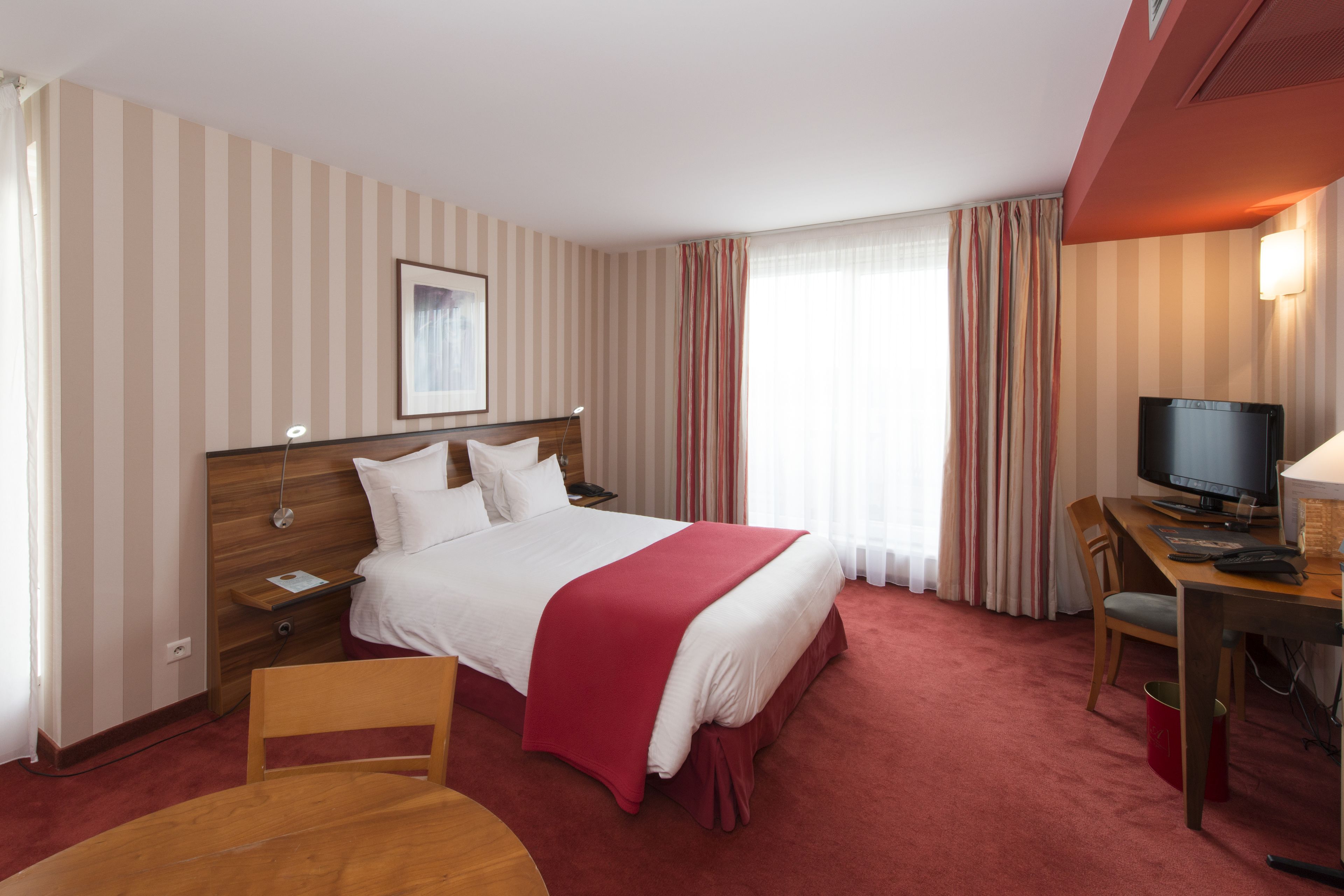 A hotel room with a bed, a desk with a computer, a chair, a TV, and a window with curtains.