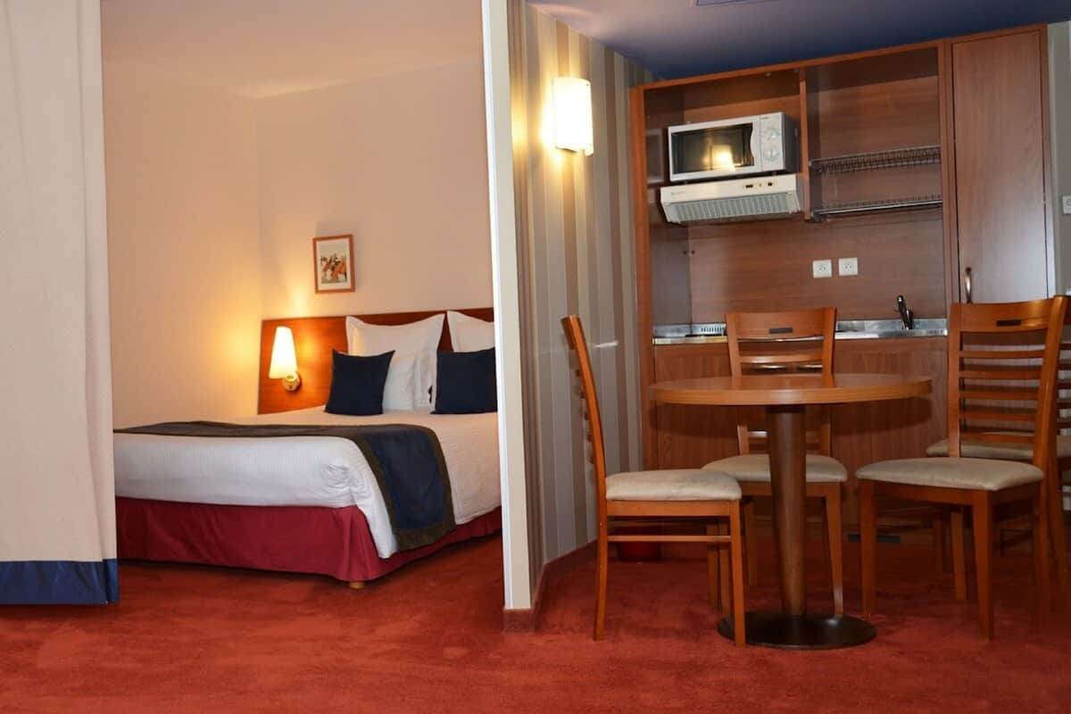 Quarto de hotel com cama, abajur e cozinha compacta com micro-ondas.