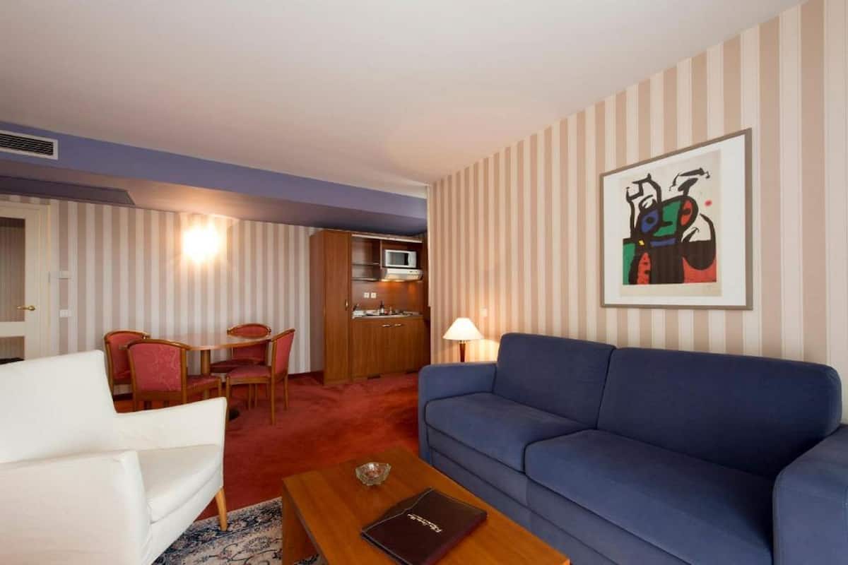 Quarto de hotel com sofá azul, poltrona branca e área de refeições com mesa e cadeiras. Possui uma kitchenette com micro-ondas e uma pintura abstrata emoldurada na parede.