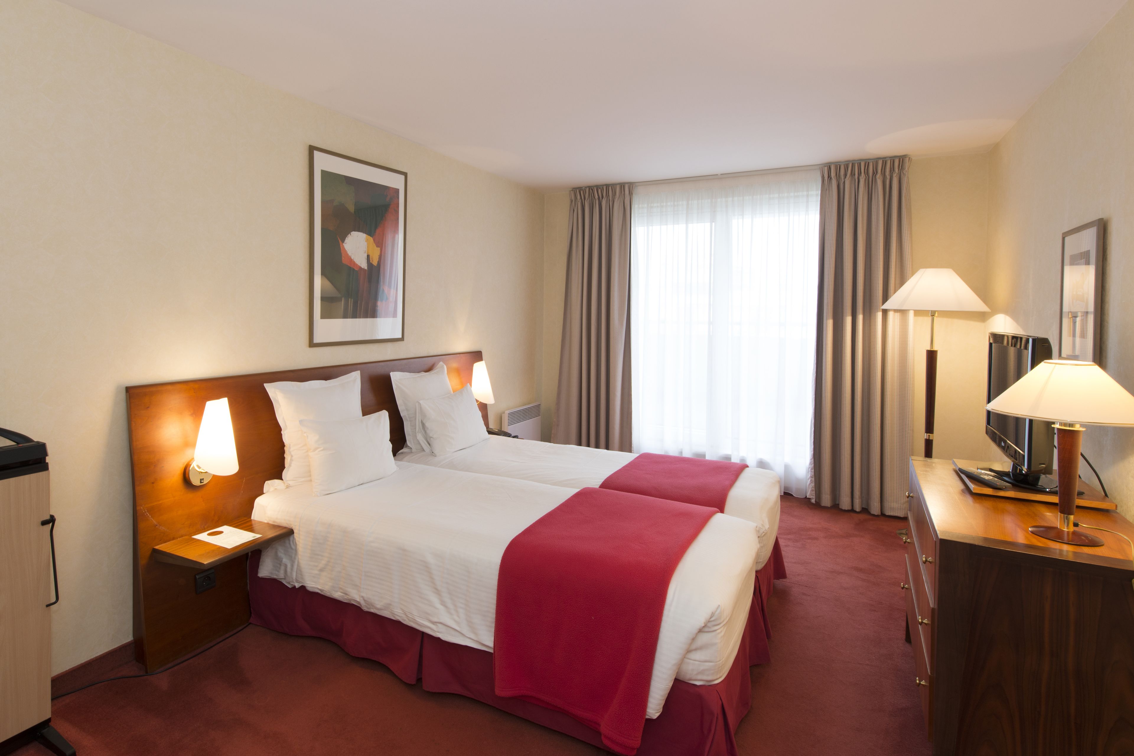 A hotel room with two beds, a desk with a lamp, a television, and a painting on the wall.