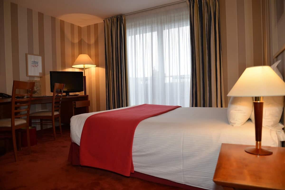 Quarto de hotel com uma cama, uma mesa de trabalho e uma televisão, e uma cadeira.