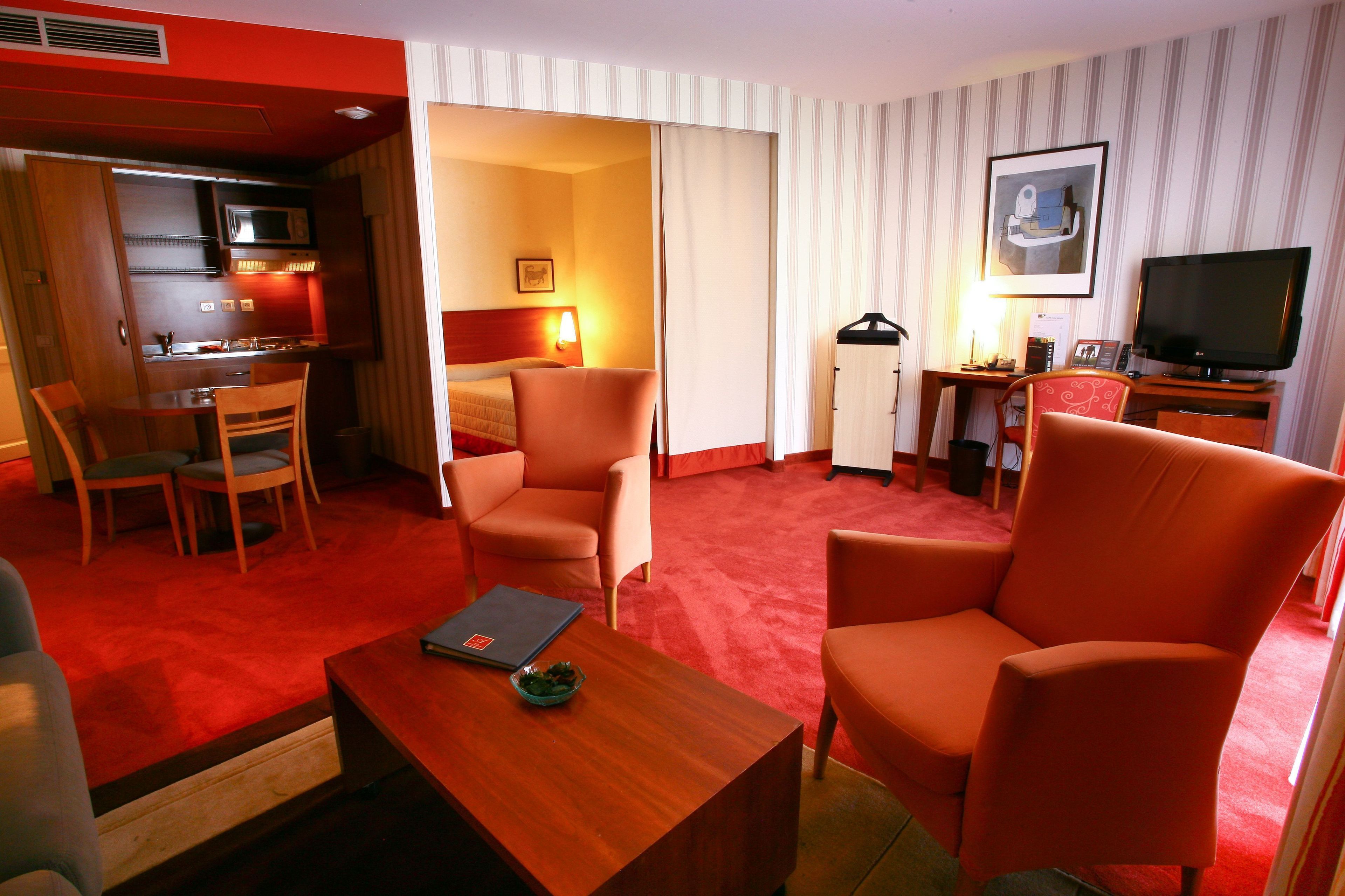 A hotel room with a kitchenette, a bed, a TV, and a seating area with a red carpet.