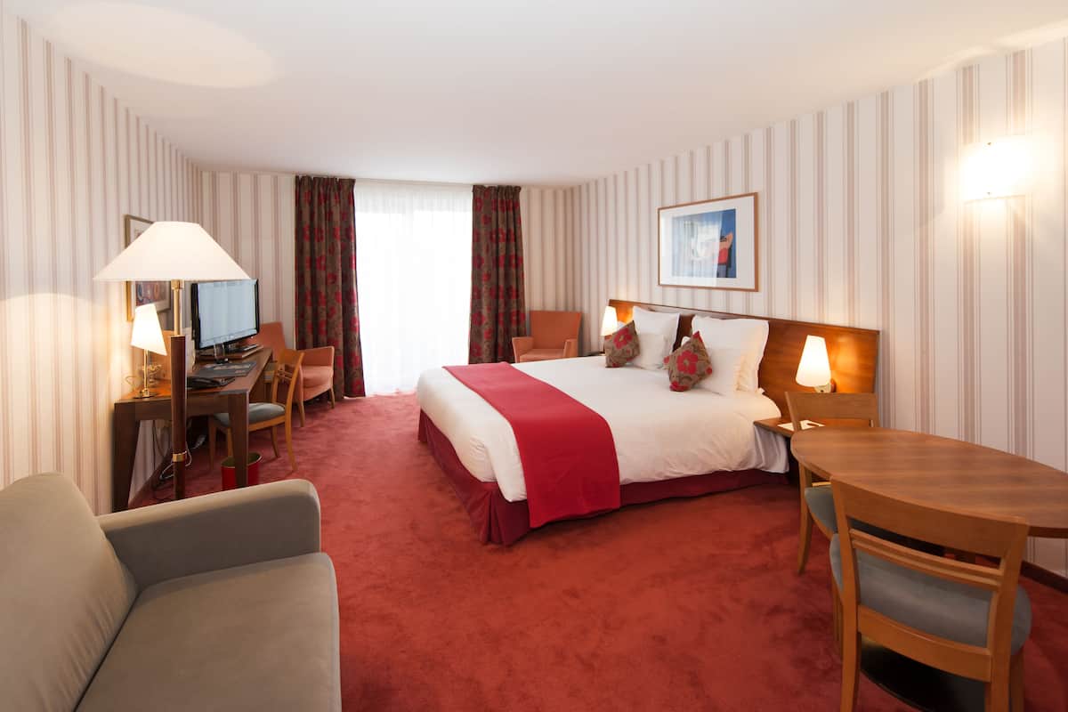 Quarto de hotel com uma cama grande, uma escrivaninha com computador, uma cadeira, uma mesa e um abajur.