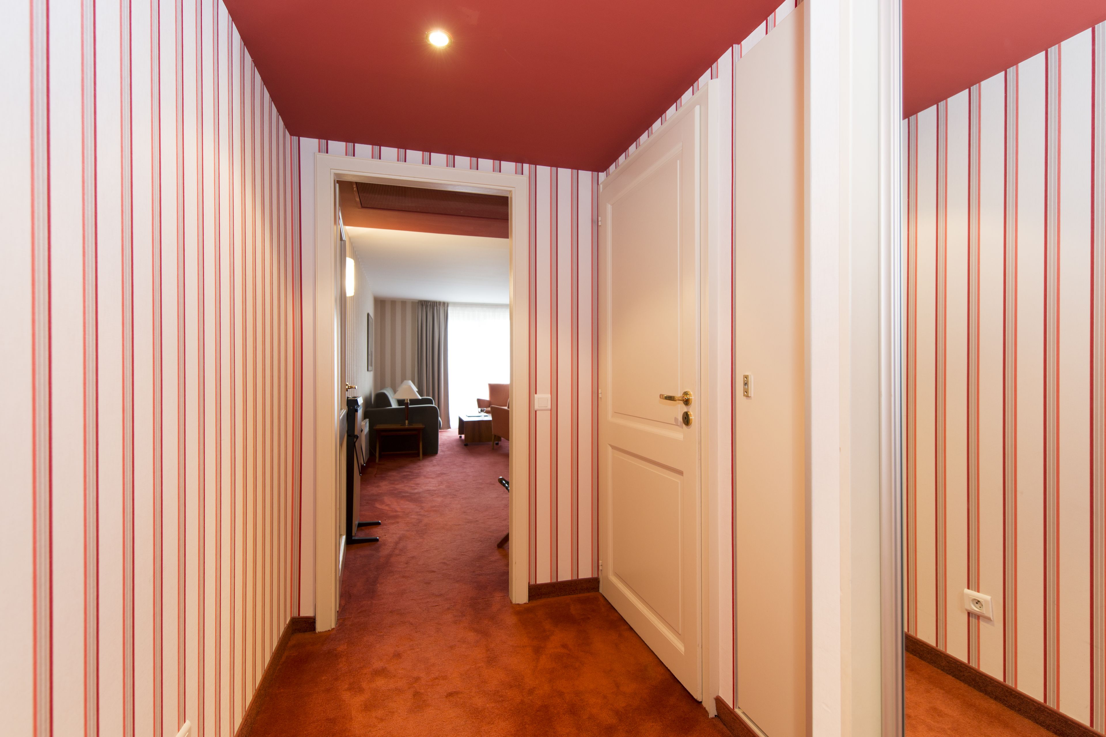 Un pasillo con papel tapiz a rayas, piso alfombrado y una puerta que da a una habitación con cama y ventana.