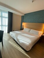 A hotel room with a large bed, a desk, a chair, a nightstand, a lamp, and a window with curtains.