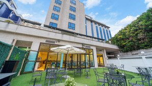Un patio de hotel con mobiliario exterior, una entrada con puerta de vidrio y un edificio alto al fondo.