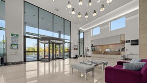Un vestíbulo amplio con entrada de vidrio, una zona de espera circular, un sofá morado y una cocina moderna al fondo.