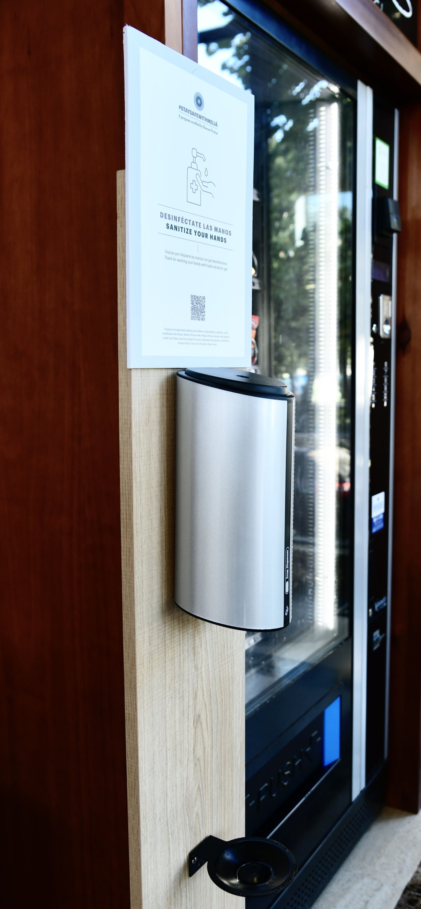 A hand sanitizer dispenser mounted on a wooden wall next to a window with a QR code and a sign.