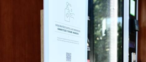 A hand sanitizer dispenser mounted on a wooden wall next to a window with a QR code and a sign.