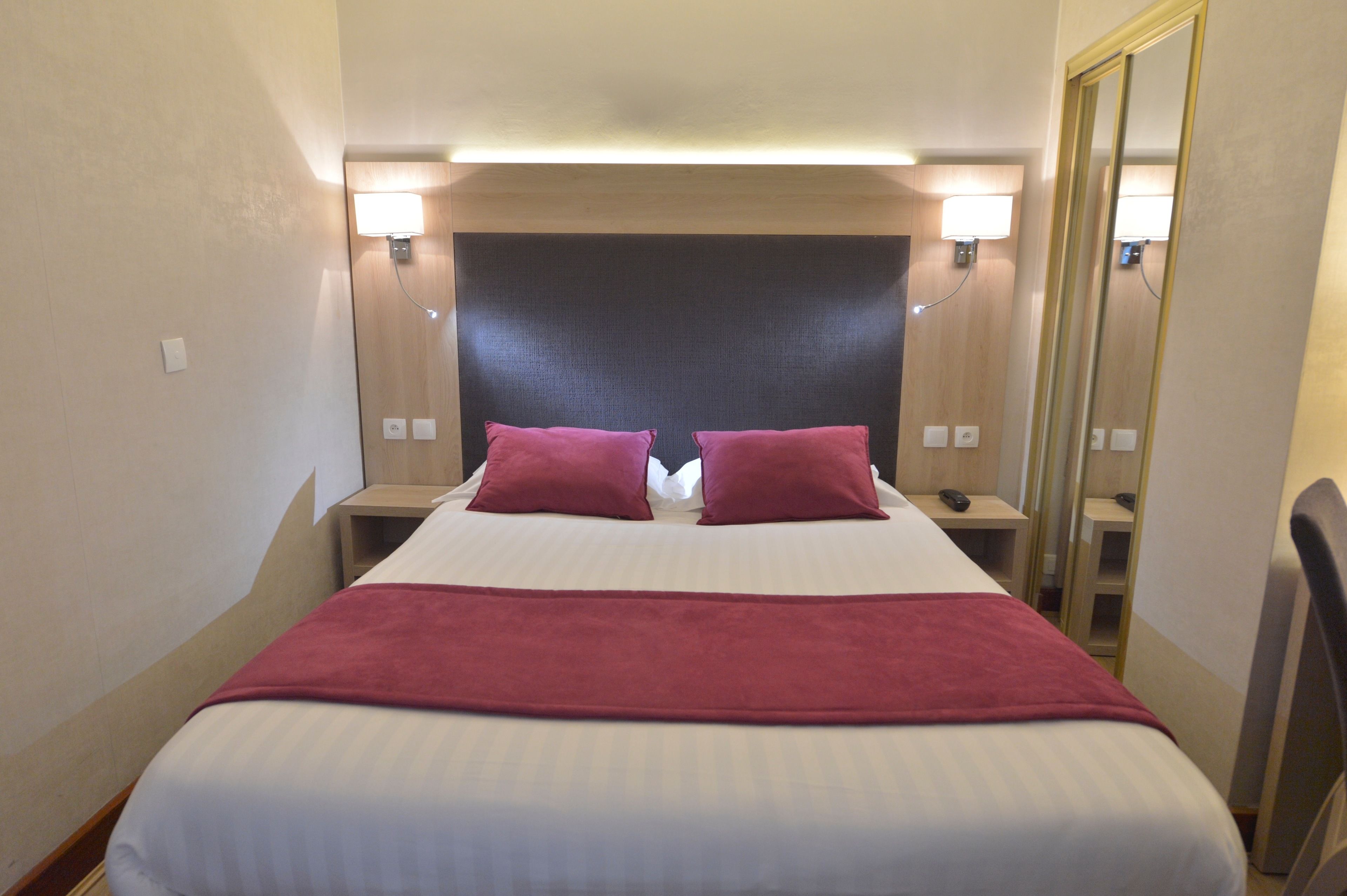 Habitación de hotel con una cama grande, dos almohadas rojas, una colcha blanca y una manta roja. Hay mesitas de noche con lámparas y un espejo en una pared.