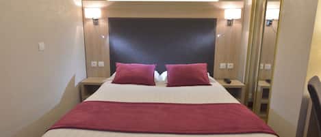 Habitación de hotel con una cama grande, dos almohadas rojas, una colcha blanca y una manta roja. Hay mesitas de noche con lámparas y un espejo en una pared.