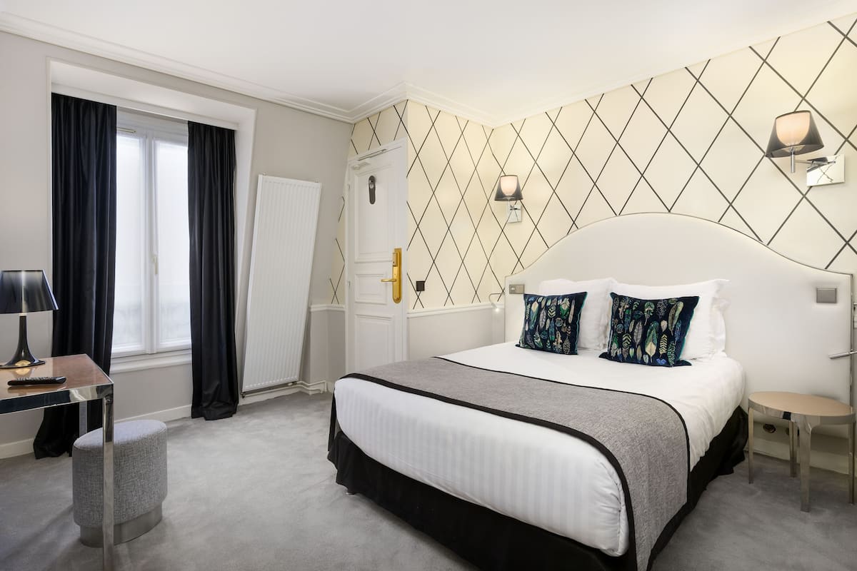 Ein Hotelzimmer mit Bett, Schreibtisch, Stuhl, einem Fenster mit Vorhängen und einer Wand mit geometrischem Muster.