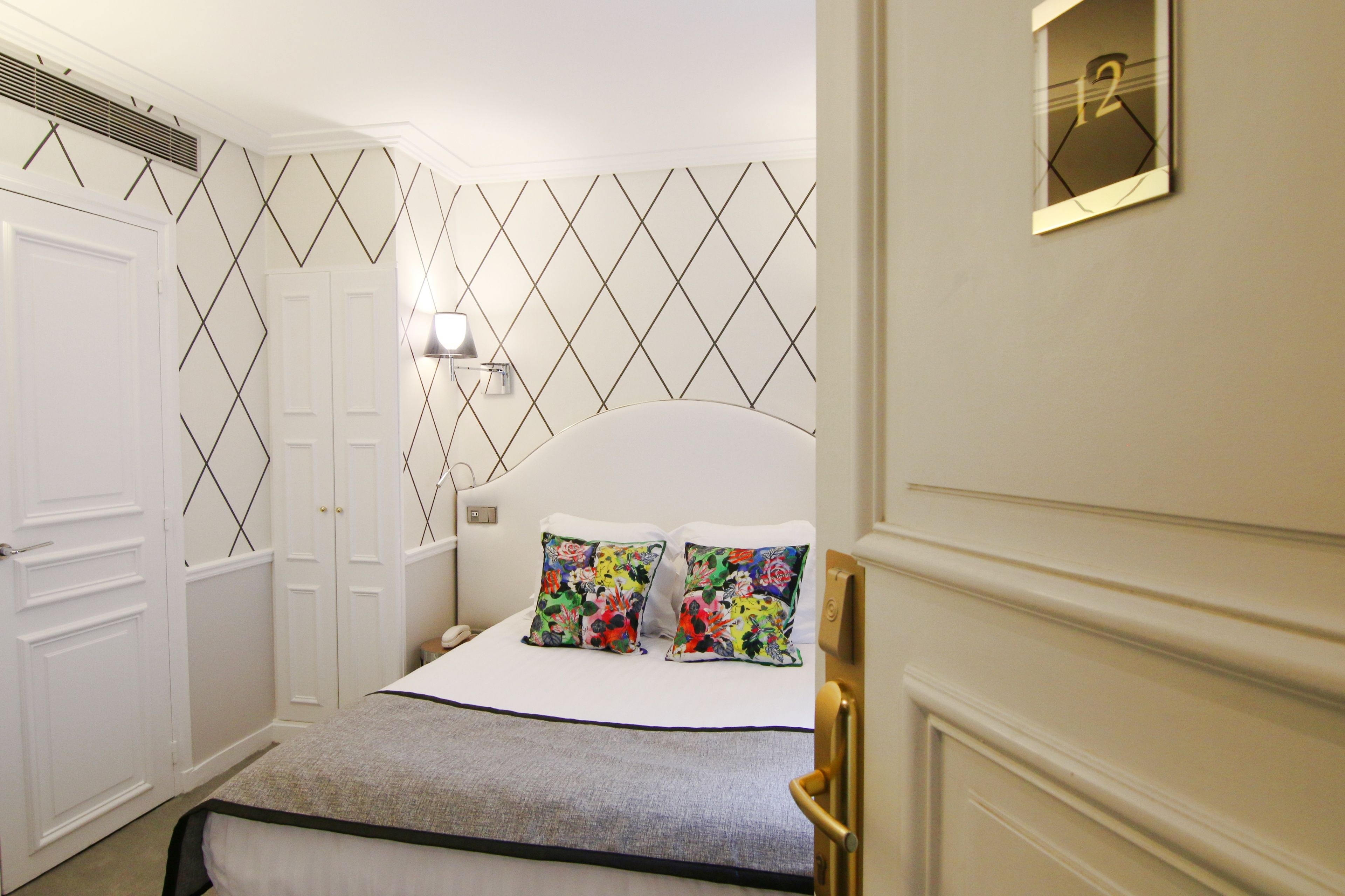 A hotel room with a bed, white walls, a geometric patterned wallpaper, a nightstand, and a wall-mounted TV.