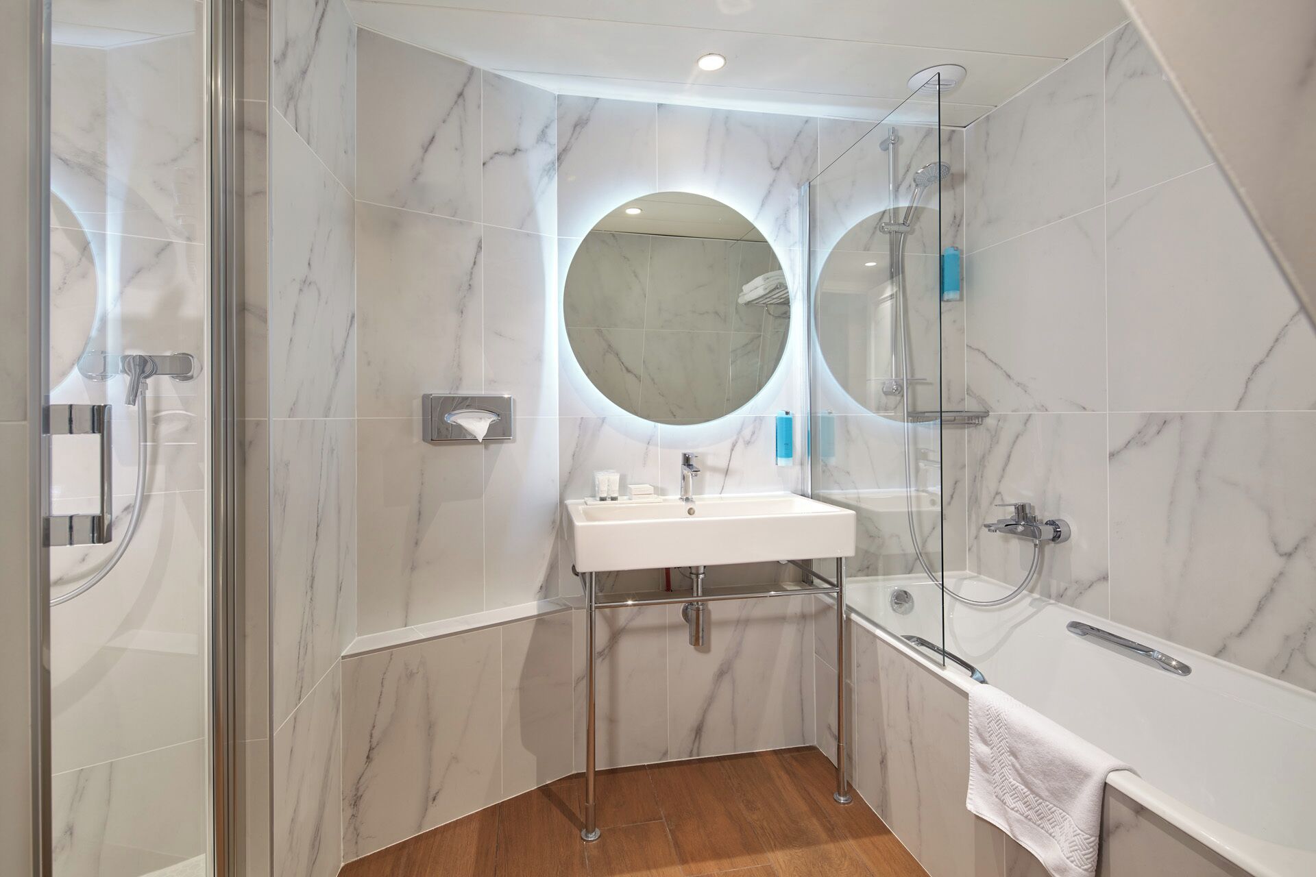 A modern bathroom with a glass-enclosed shower, a round mirror, a white sink, and a wooden floor.