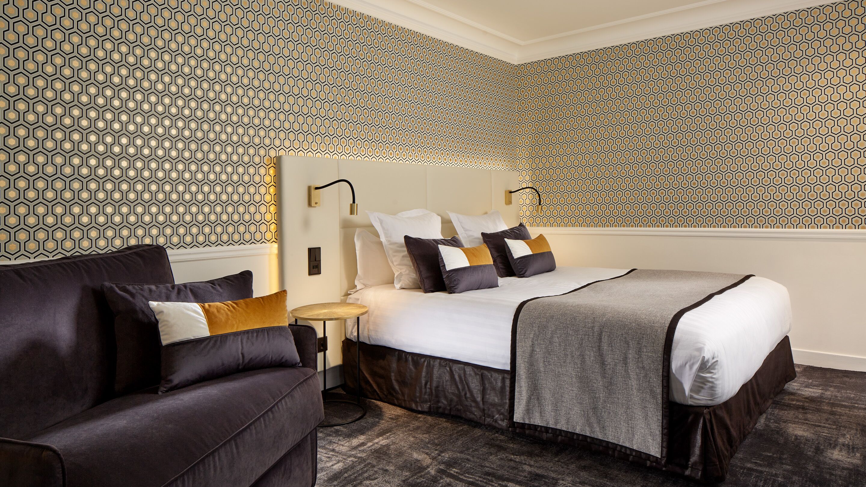 Ein Hotelzimmer mit gemustertem Wandteppich, einem Bett mit vielen Kissen, einem dunkelgrauen Sofa und einem kleinen Nachttisch mit Lampe.