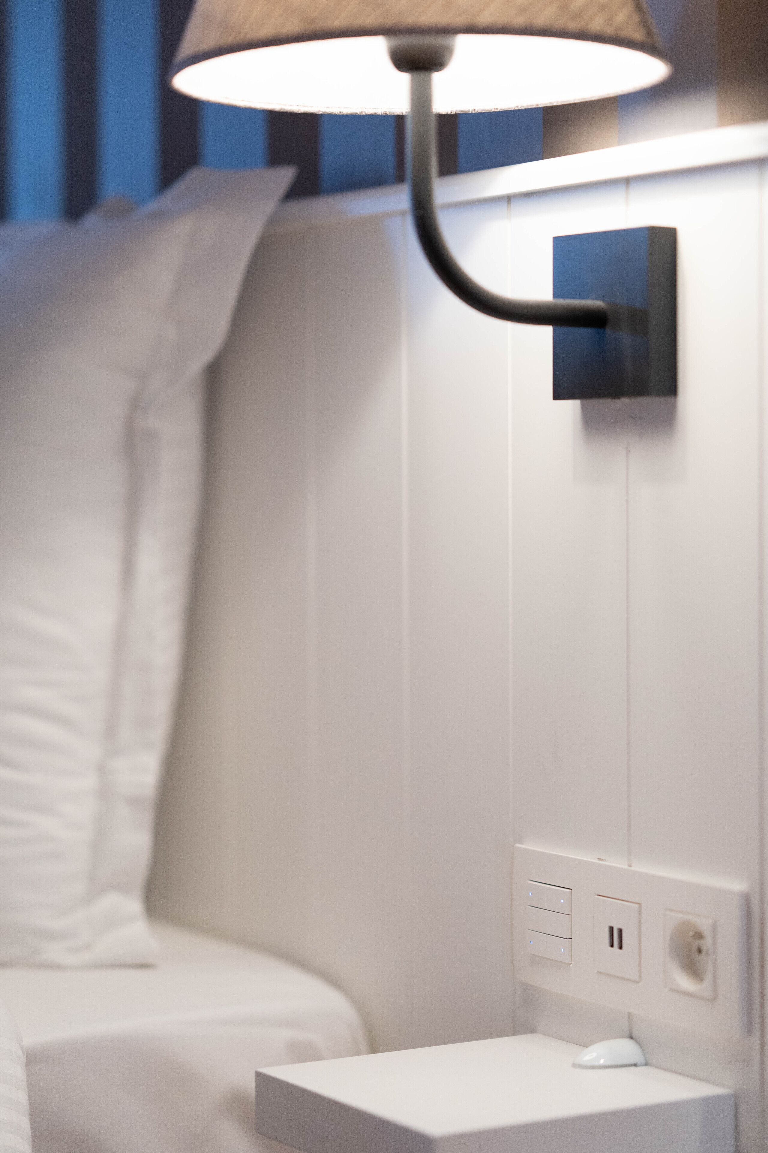 Un dormitorio moderno con una cama blanca, una mesita de noche, una lámpara y un enchufe en la pared.