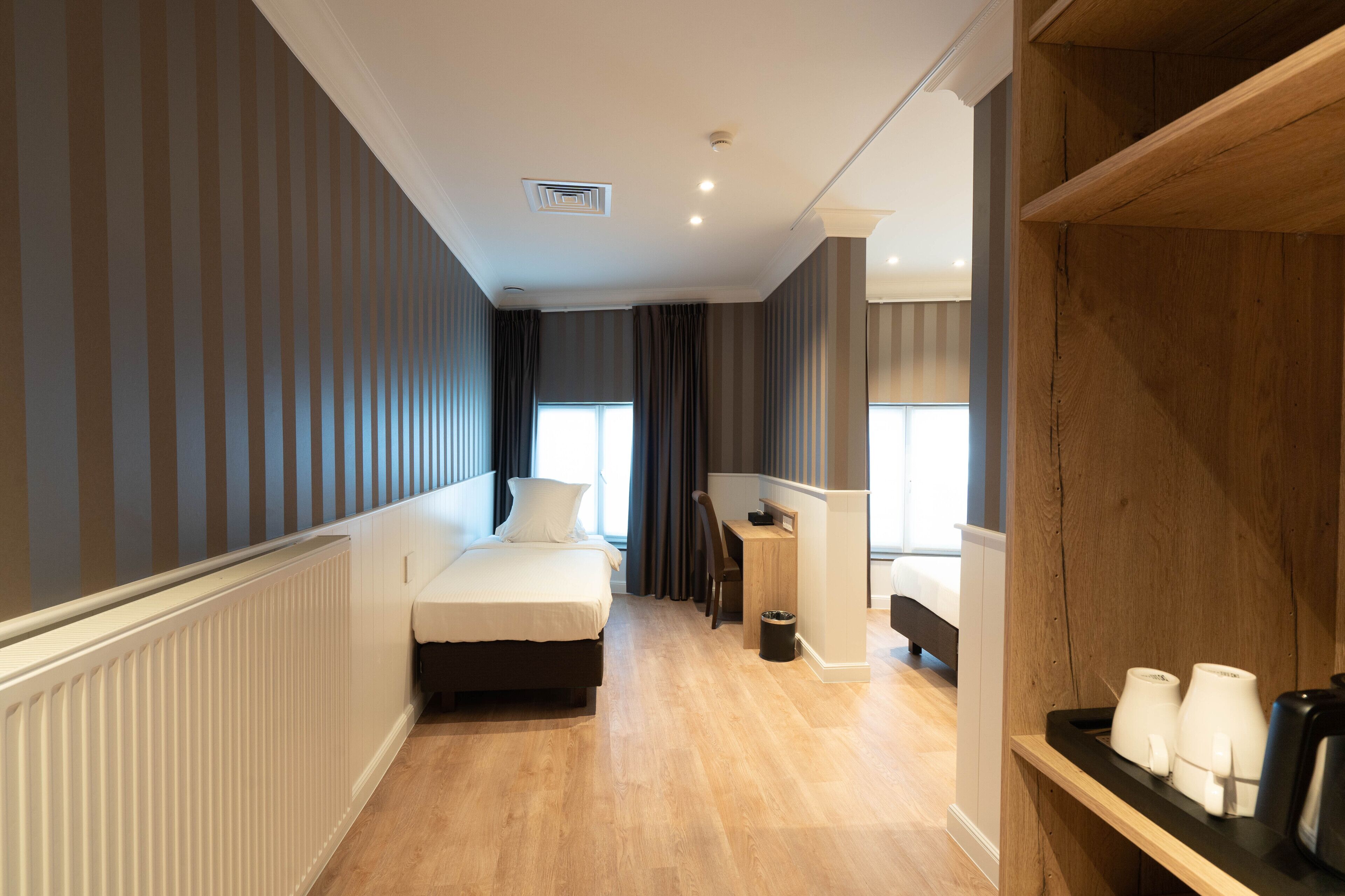 A hotel room with a bed, a desk, a chair, a radiator, and a shelf with cups.