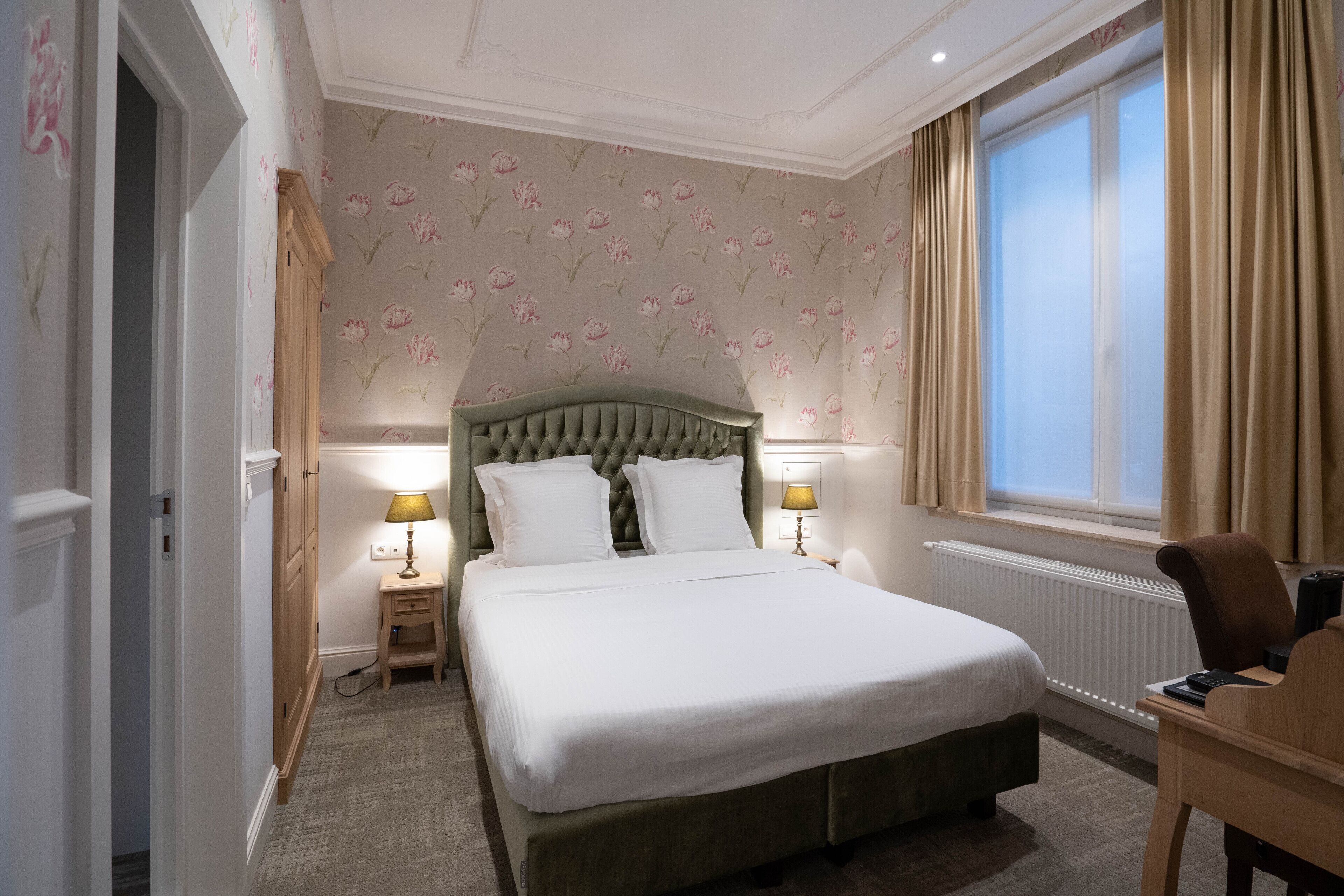 A neatly made bed with white linens, a wooden nightstand with a lamp, and a window with curtains.