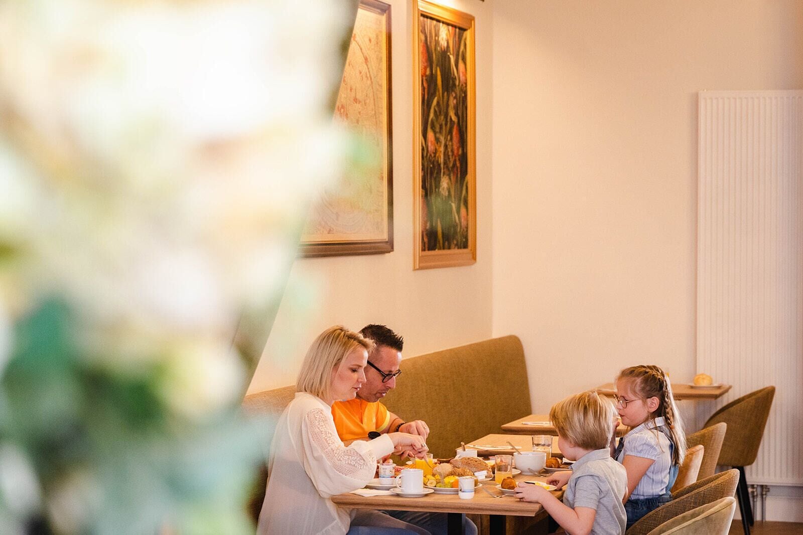 A family dining at a table with breakfast items, in a room with a radiator and framed artwork on the wall.