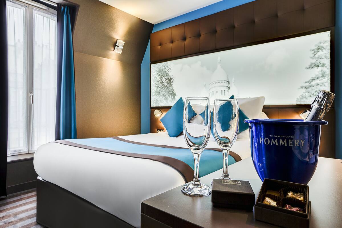 Habitación de hotel con cama, mesita de noche con cubo para champán, dos copas y una caja de bombones.