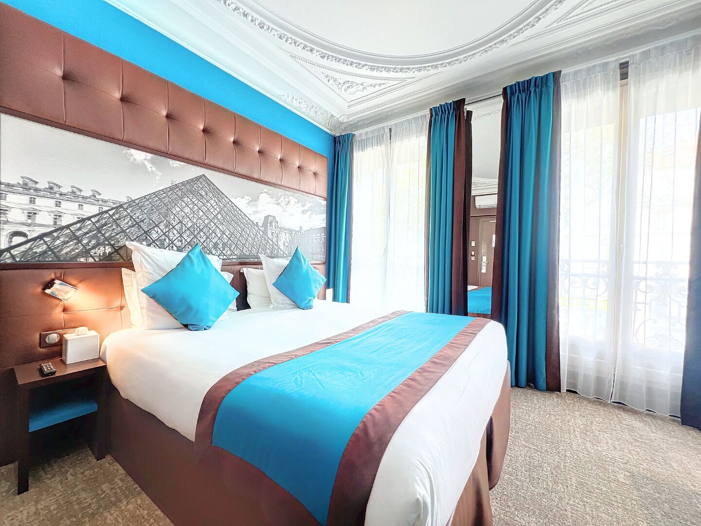 A hotel room with a large bed, a headboard with a mural, a nightstand with a lamp, and a view of the Eiffel Tower through the window.