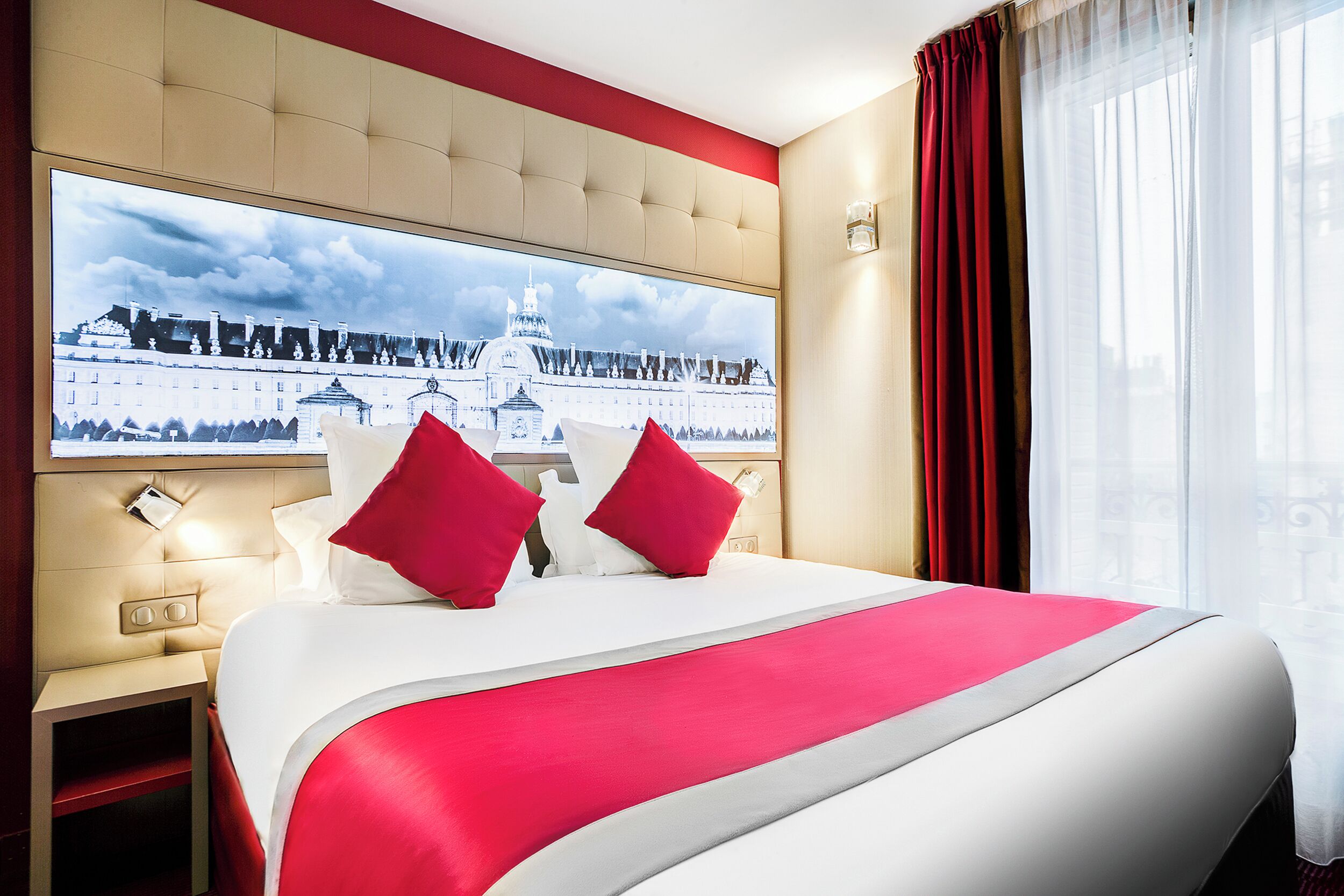 A hotel room with a large bed, red and white bedding, a view of a cityscape, and a red curtain.