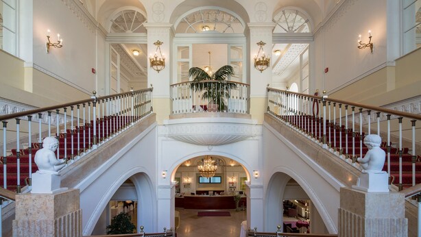 Een statig hotel ontvangsthal met een centrale trap, weelderige kroonluchters en klassieke beelden.