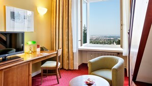 Hotelkamer met bureau, stoel, televisie, lamp en uitzicht op een stad door een raam.