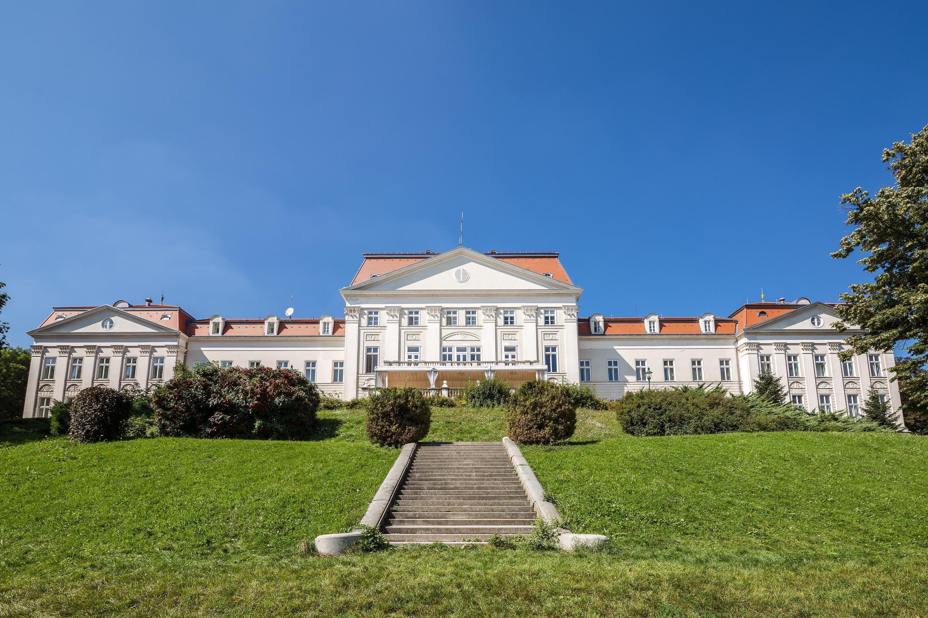A large, symmetrical building with a central entrance and a red roof, surrounded by a well-maintained lawn and bushes.
