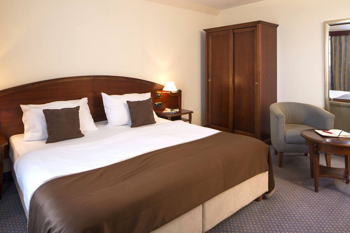 Habitación de hotel con una cama grande, un armario, una silla y una mesita de noche con lámpara.