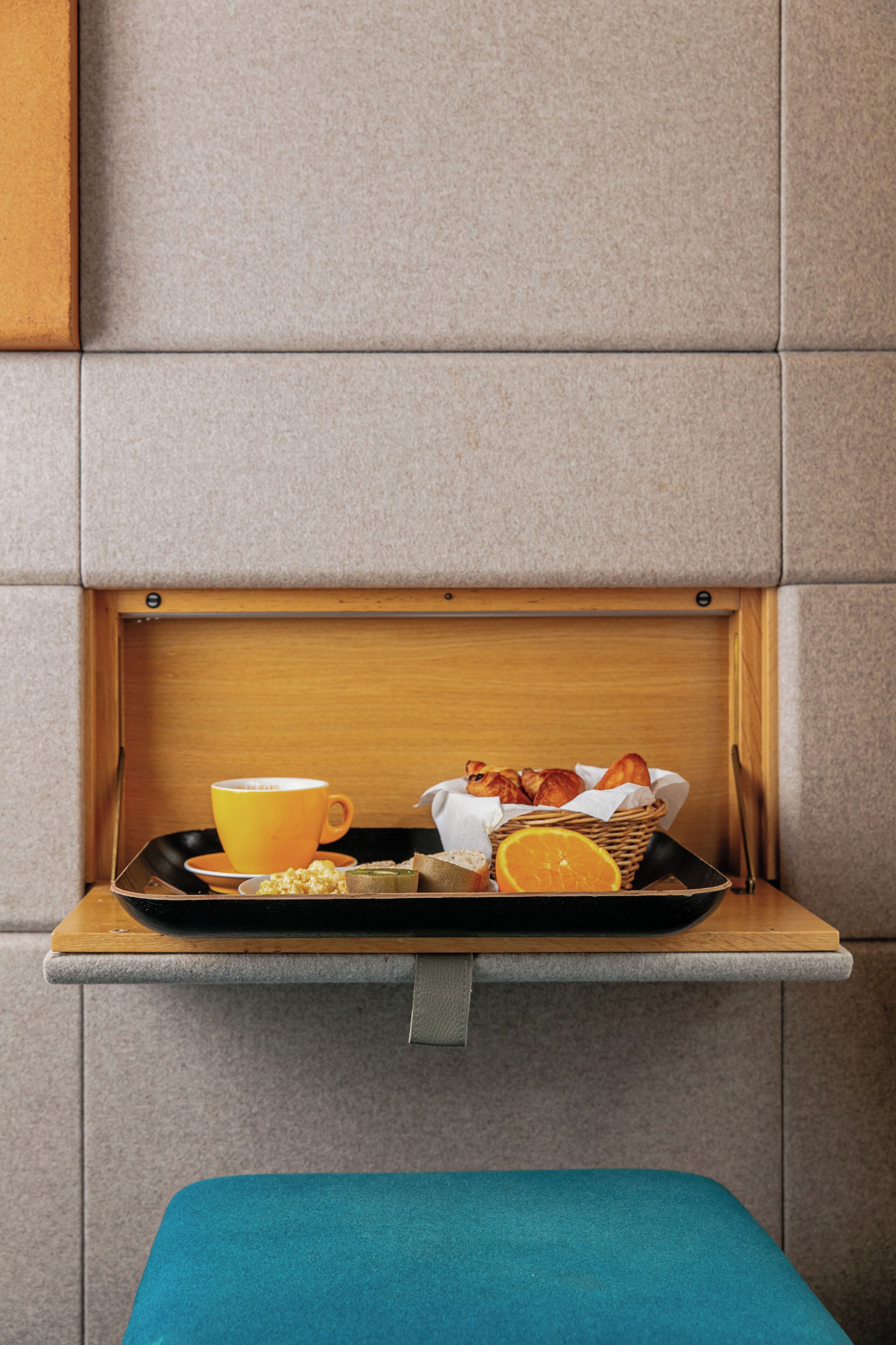 Bandeja de desayuno con una taza, pasteles y una naranja, sobre una bandeja en una habitación con pared gris y repisa de madera.