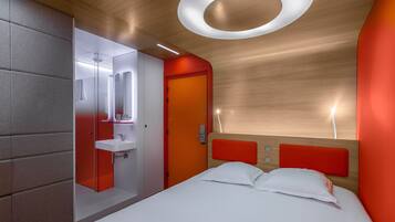 Una habitación de hotel moderna con techo de madera, pared de acento roja, cama blanca y baño con espejo y lavamanos.