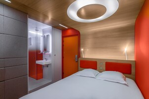 Una habitación de hotel moderna con techo de madera, pared de acento roja, cama blanca y baño con espejo y lavamanos.