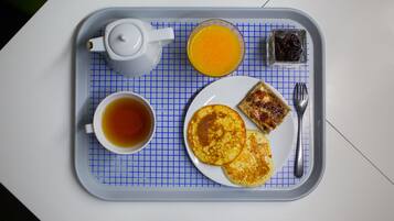 Tarjottimella on teekannu, teekuppi, lautanen pannukakkuja, lasi appelsiinimehua ja pieni purkki hilloa.