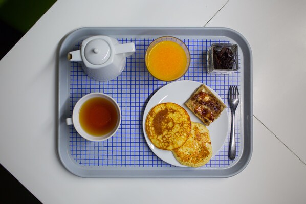 A tray with a teapot, cup of tea, plate of pancakes, glass of orange juice, and a small container of jam.
