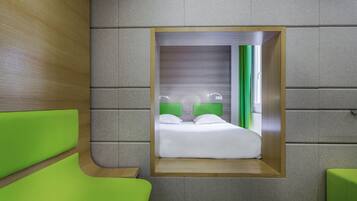 Ein kleines Hotelzimmer mit einem fest eingebauten Bett, grünem Polstersitzmöbel und einer Wand in neutralen Farbtönen.