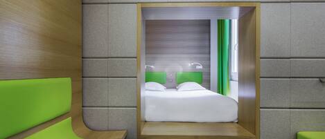A compact hotel room with a built-in bed, green upholstered seating, and a neutral-toned wall.