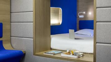 Una habitación de hotel moderna con una cama, una mesita de noche con un libro, una bandeja con alimentos para el desayuno y una lámpara en la mesita de noche.