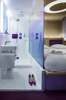 A modern hotel room with a glass-enclosed shower, a white sink, a bedside table with a tray of breakfast items, and a neatly made bed.