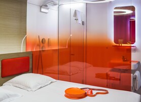 A modern hotel room with a bed, a red headboard, a glass partition, a shower area, and a bathroom with a mirror and towel rack.