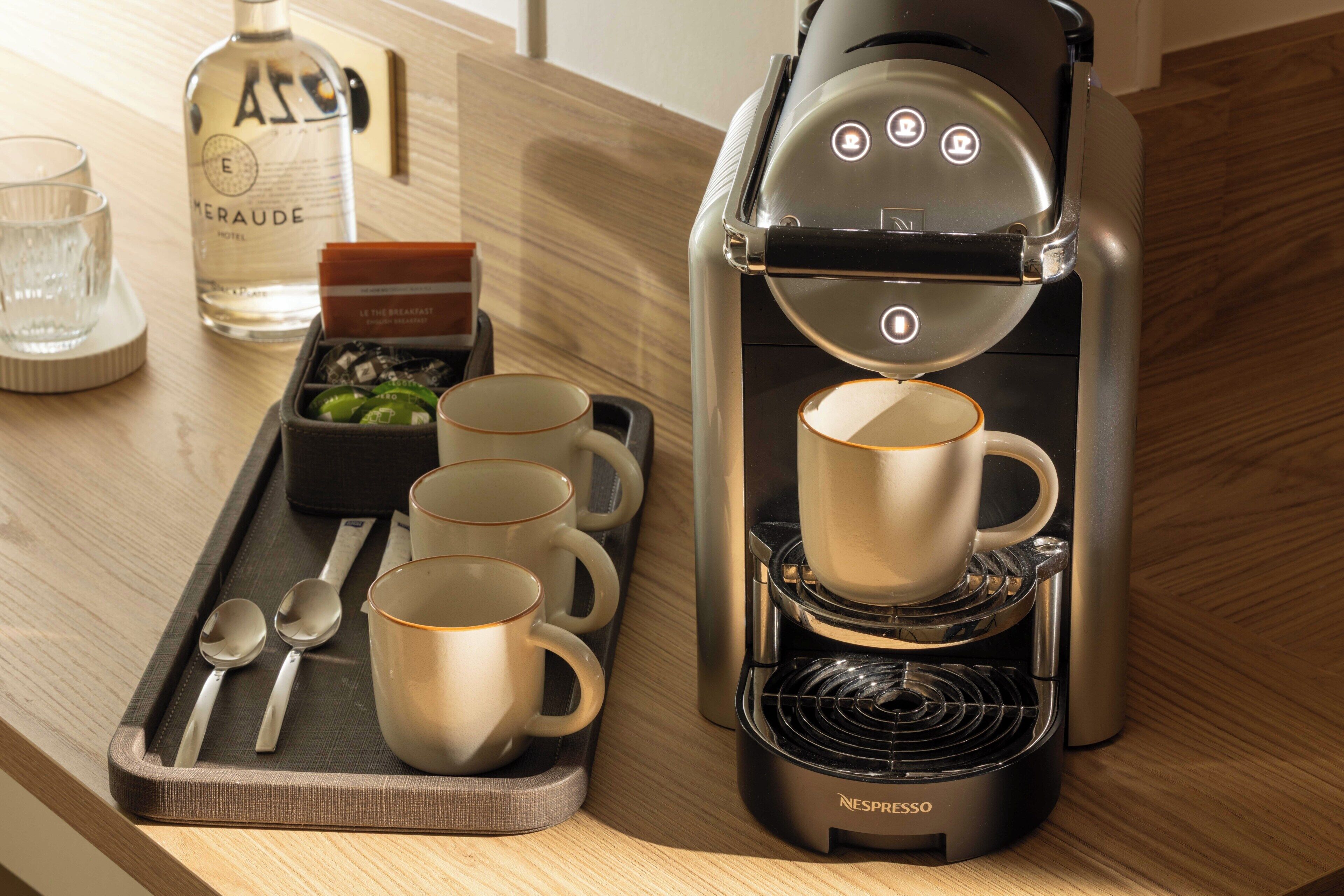 Uma máquina de café Nespresso em uma bancada de madeira, com uma bandeja contendo quatro xícaras, duas colheres e uma garrafa de água.