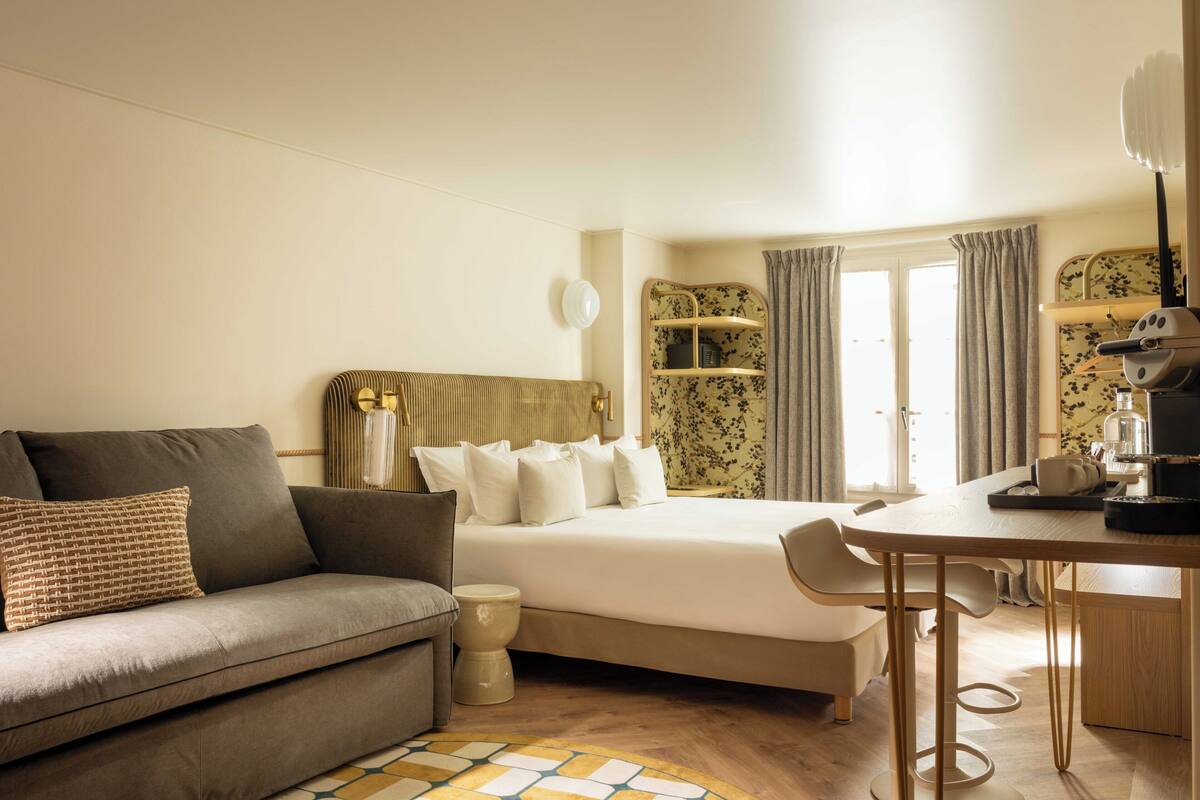 A bedroom with a bed, a sofa, a bedside table, a lamp, a coffee machine, and a window with curtains.
