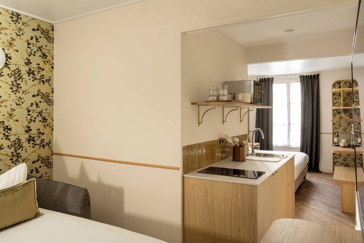 A compact hotel room with a kitchenette, a bed, a bedside table, a chair, and a window with curtains.