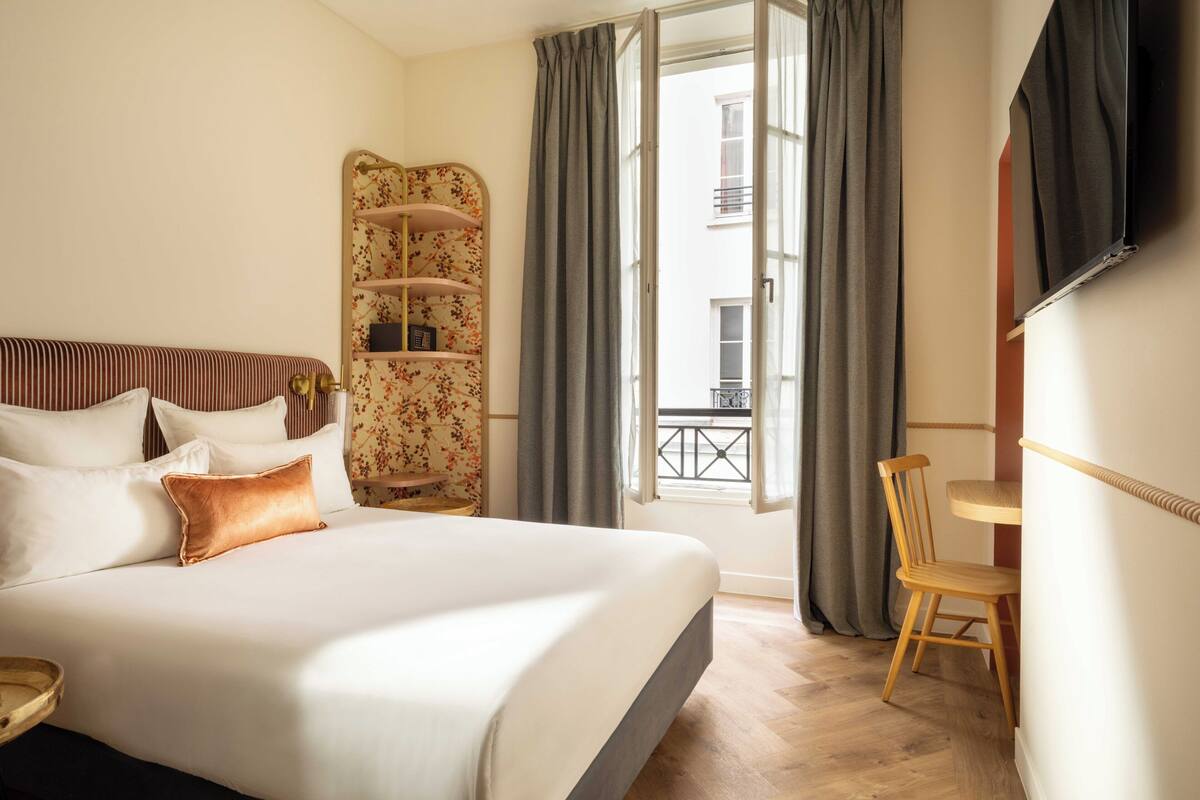 A hotel room with a bed, a wooden chair, a bookshelf, and a balcony with curtains.