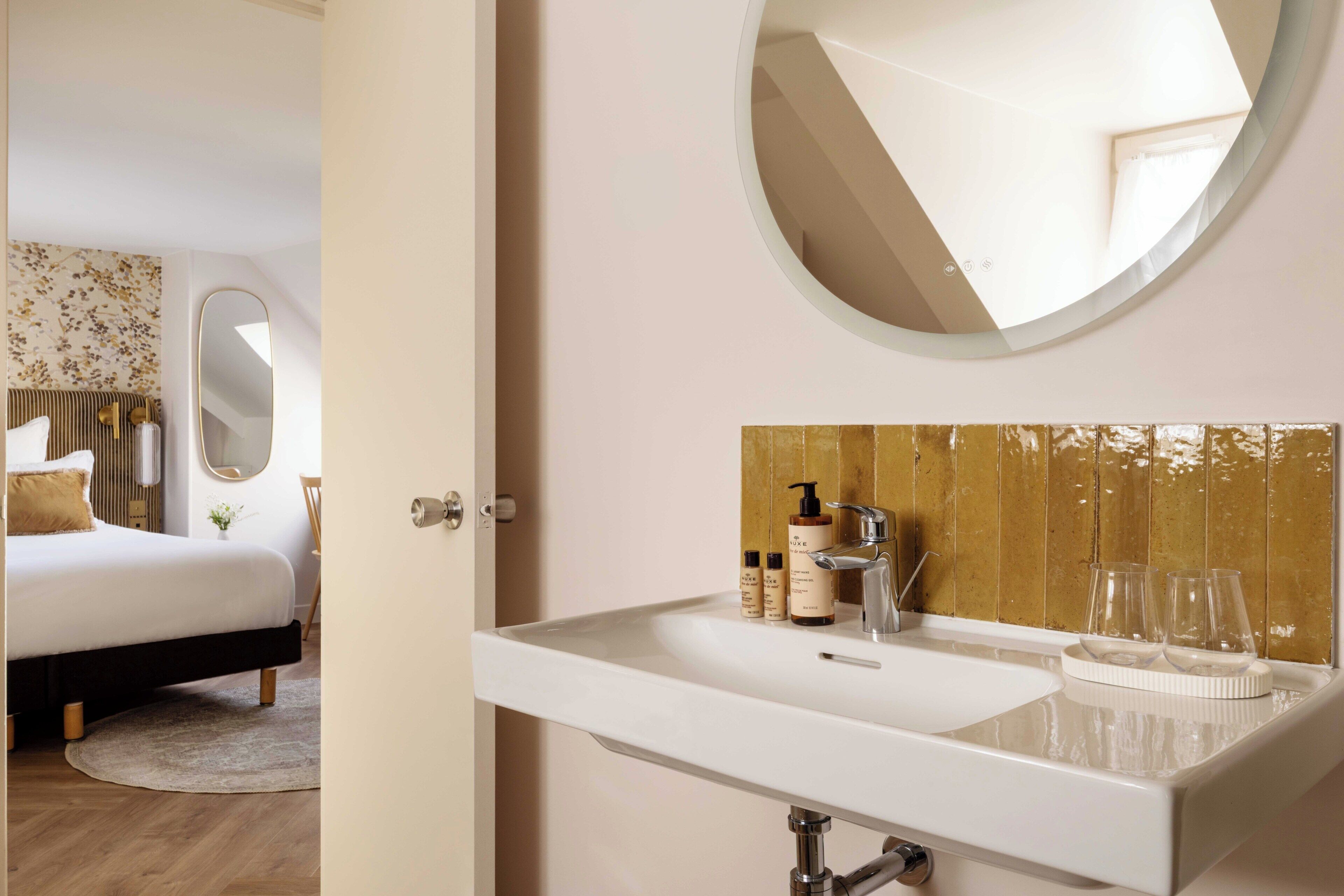 A hotel room with a bed, a round mirror, a sink with toiletries, and a view of a wooden wall.