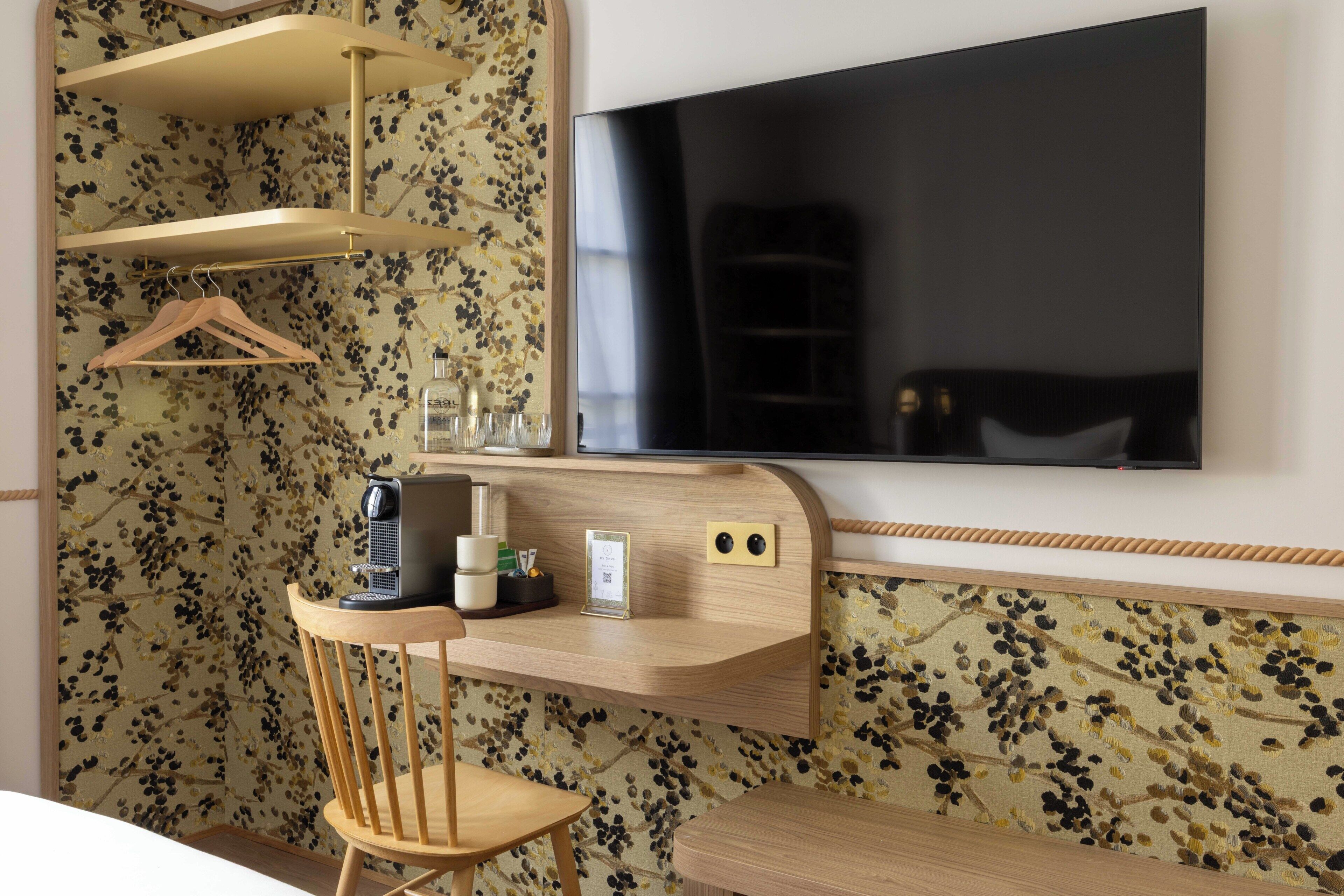 Une chambre d’hôtel avec un bureau en bois, une chaise, un téléviseur fixé au mur et un papier peint à motifs floraux.